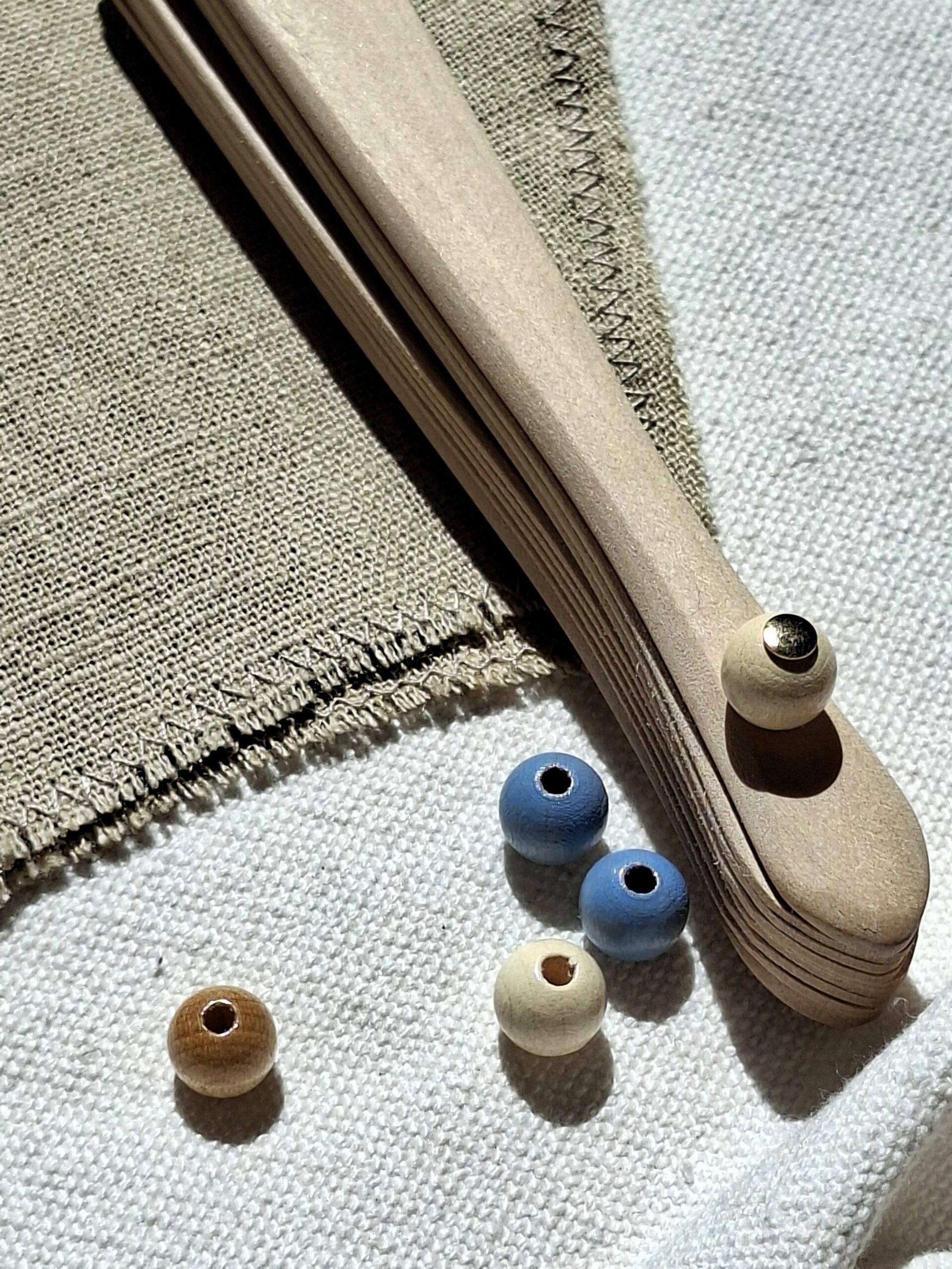 Un primer plano de componentes para la fabricación de un abanico de lino artesanal, que muestra varillas de madera curvada apiladas, varias cuentas de madera (azules, naturales y una con herraje dorado) y retales de tela de lino rústico sobre un fondo de canvas blanco, con luz natural directa.