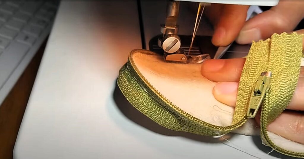 Un primer plano de primer plano de manos cosiendo una cremallera verde oliva en el borde de un monedero gordito de fieltro de lana 100% natural de color crema, con una máquina de coser, mostrando la aguja y el pie prensatelas en acción, sobre un fondo de costura desenfocado. ARQ2eme