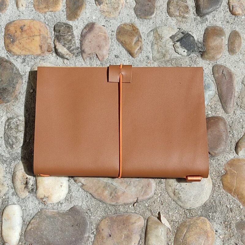 Nosotras 2 Un cuaderno de piel camel cerrado con un cierre elástico de color naranja y detalle metálico naranja sobre un fondo de un suelo de mosaico de piedras