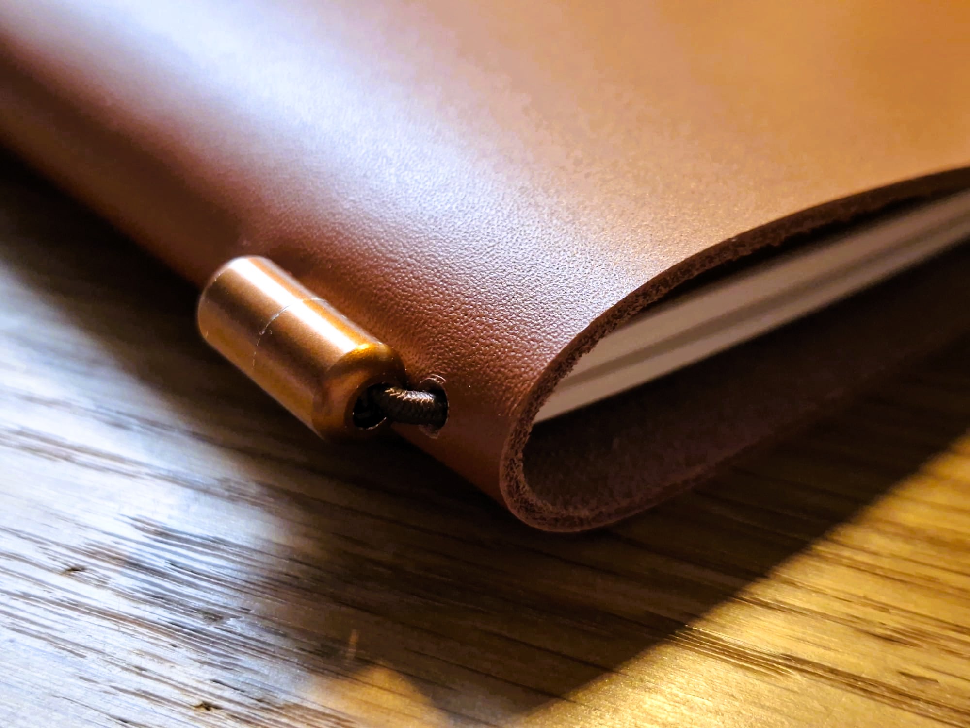 Primer plano de herraje metálico cilíndrico en tono bronce sobre el lomo de un cuaderno de piel vacuna color camel, destacando la textura natural del cuero y el sistema de cierre elástico.