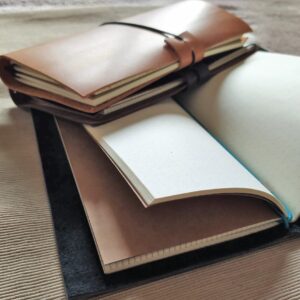 Un conjunto de cuadernos de piel artesanales apilados sobre fondo texturizado, que muestra un cuaderno de piel camel sobre uno marrón oscuro, y un cuaderno de piel negro abierto revelando recambios de papel blanco con cuadrícula de puntos y un cierre elástico azul.